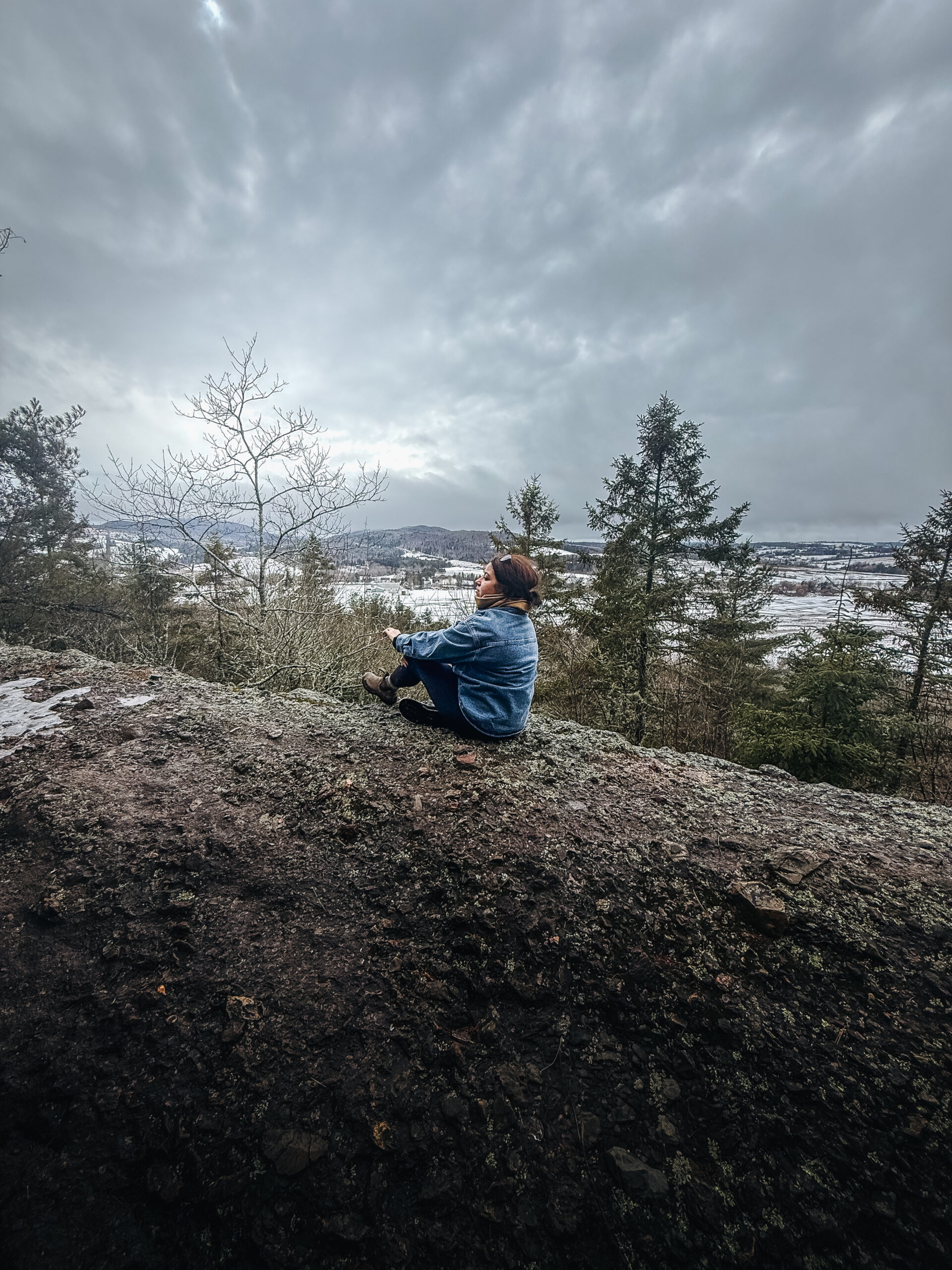 Woman sitting on a snow-covered rocky bluff overlooking the Sussex valley in New Brunswick in winter