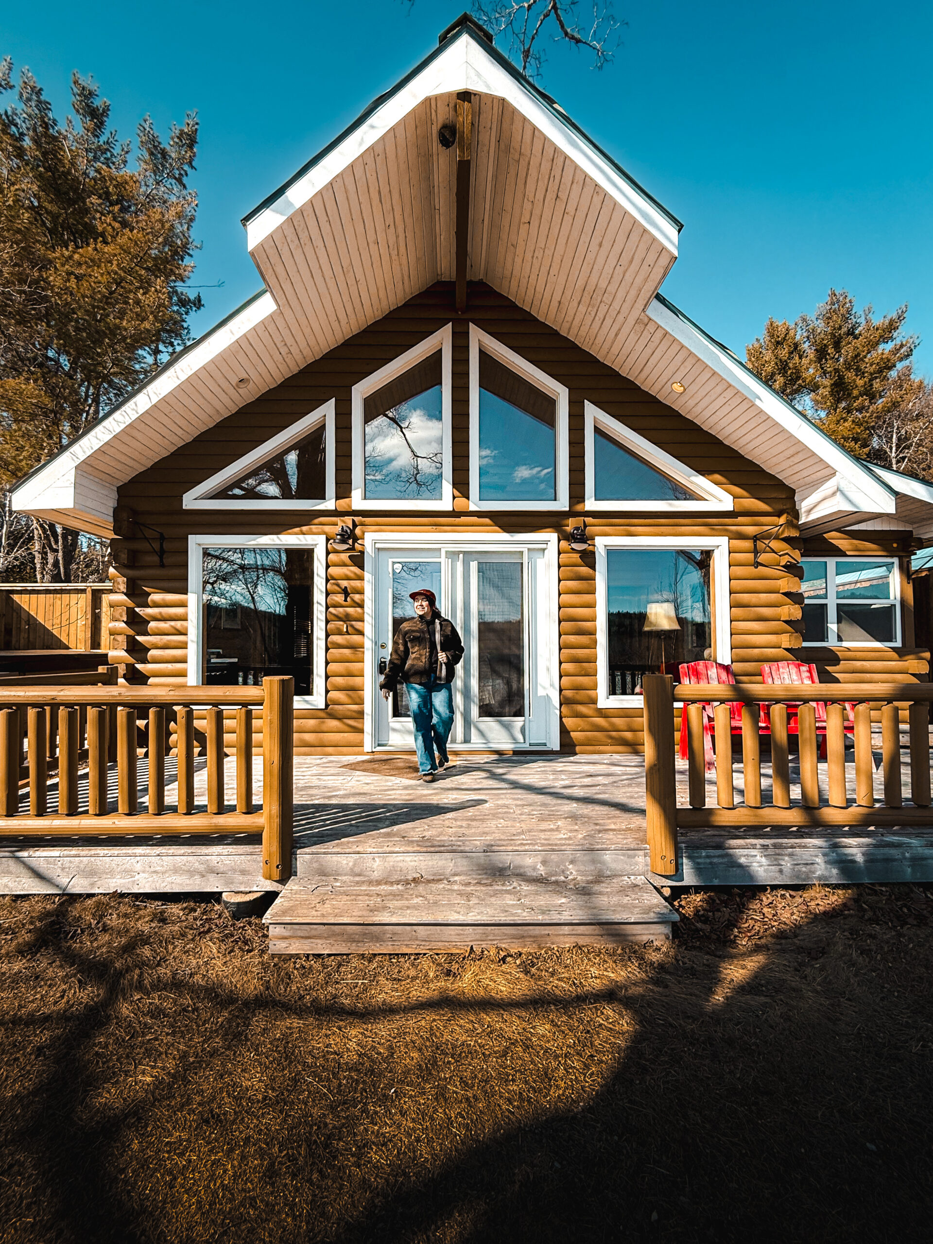 Woman walking out the front door of a log cabin at Old River Lodge in Boiestown New Brunswick with red Adirondack chairs on the deck and A-frame windows under a blue sky