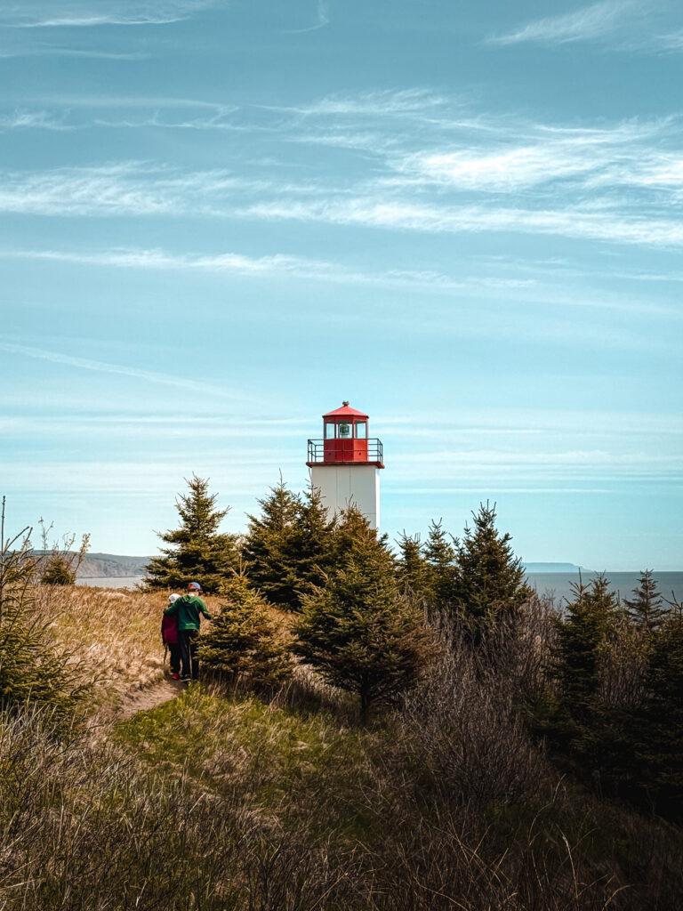 Two hikers walking toward the red and white Quaco Head Lighthouse Trail landmark through spruce trees and coastal grass in West Quaco NB