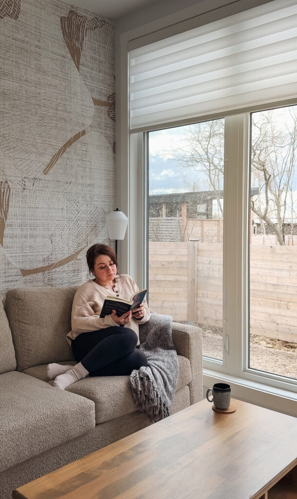 Melissa reading a book on a couch inside a Glamping Aboiteau unit with warm morning light coming through large windows and a coffee mug on the table