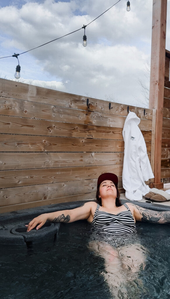 Woman relaxing with eyes closed in a private hot tub at Glamping Aboiteau with string lights overhead and a white robe hanging nearby