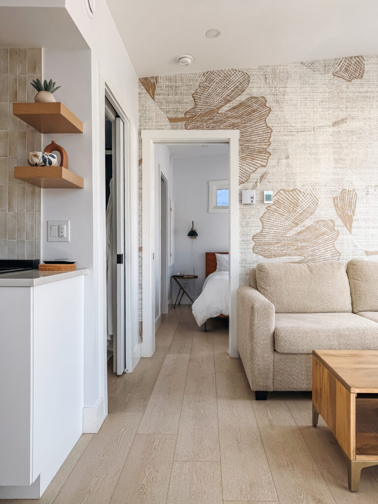 Open-plan living area of a Glamping Aboiteau unit with botanical wallpaper couch and a view through the hallway into the bedroom