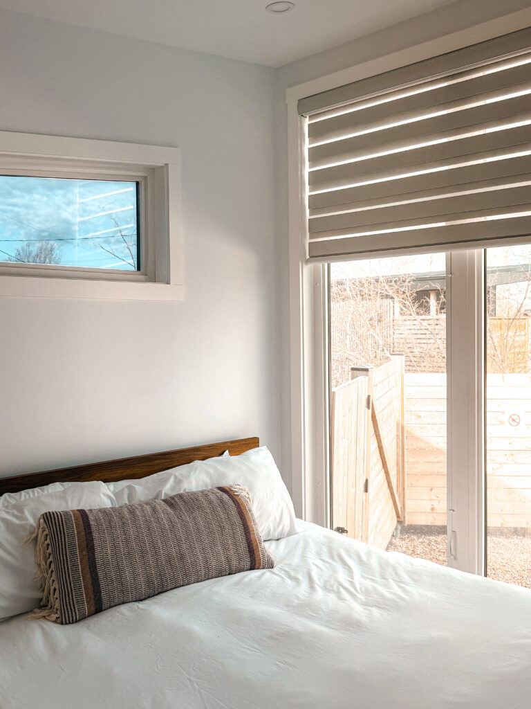 Cozy bedroom inside a Glamping Aboiteau unit with white linen bedding a wood headboard and a sliding glass door to the private yard
