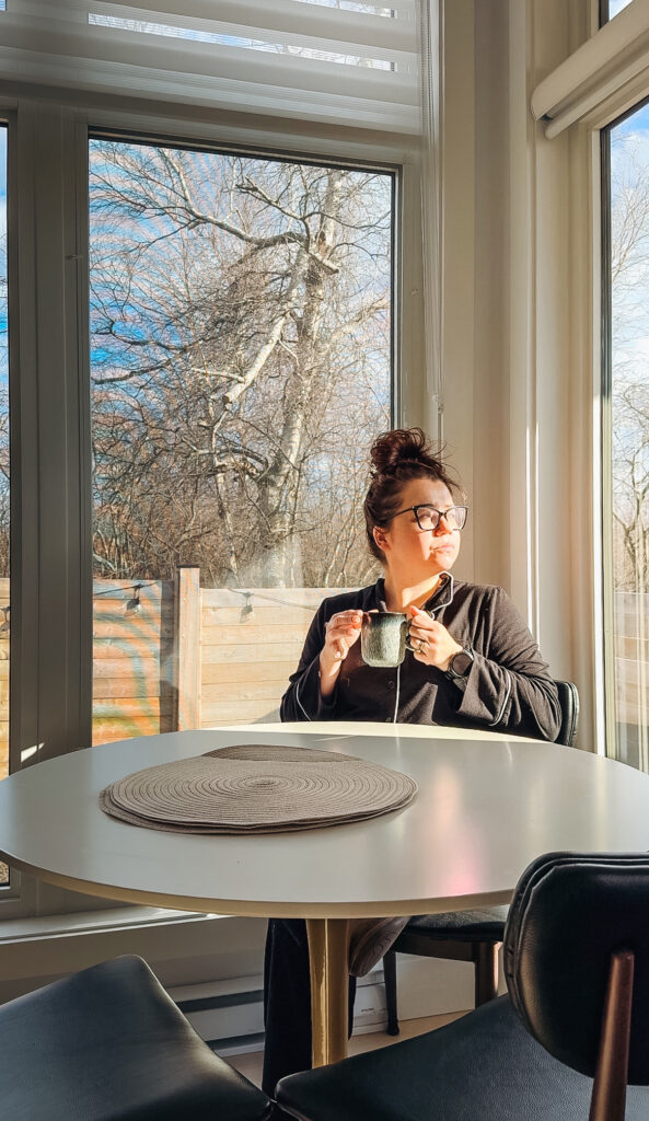 Woman sitting at a round dining table holding a mug inside a Glamping Aboiteau unit with soft morning light through large windows