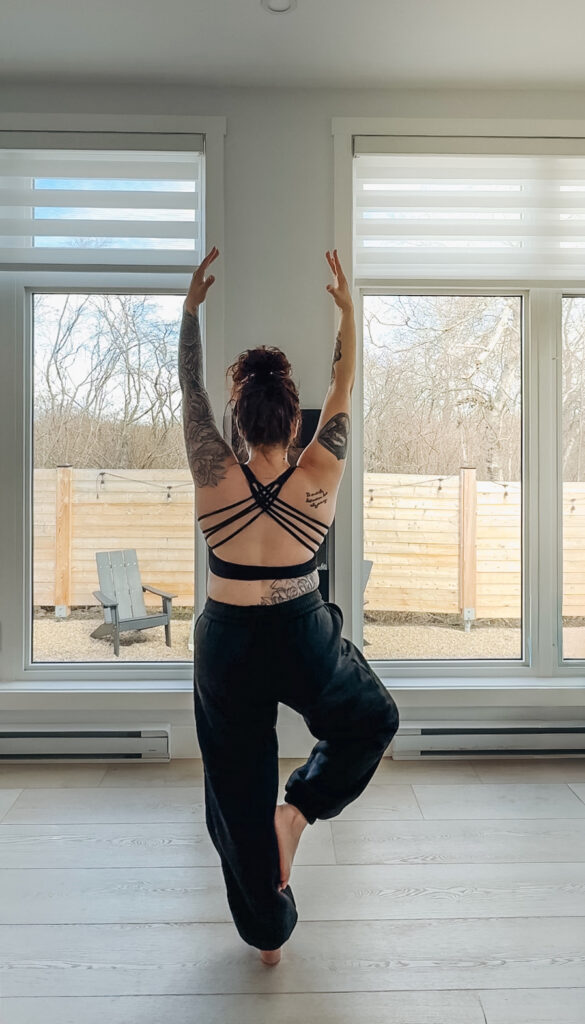 Woman doing a yoga tree pose inside a glamping unit at Glamping Aboiteau with large windows overlooking the private yard