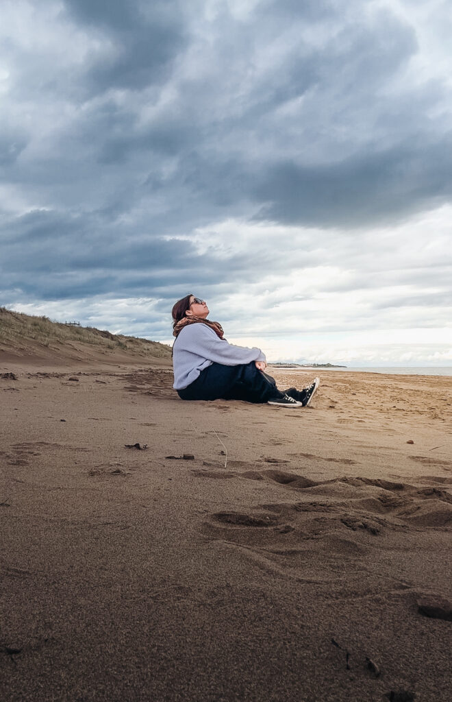 Woman sitting alone on Aboiteau Beach in Cap-Pelé New Brunswick under a wide dramatic cloudy sky