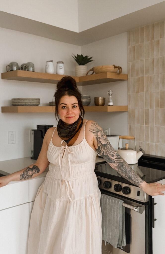 Melissa Danielle in the full kitchen of a Glamping Aboiteau unit showing open shelving and a ceramic tea kettle