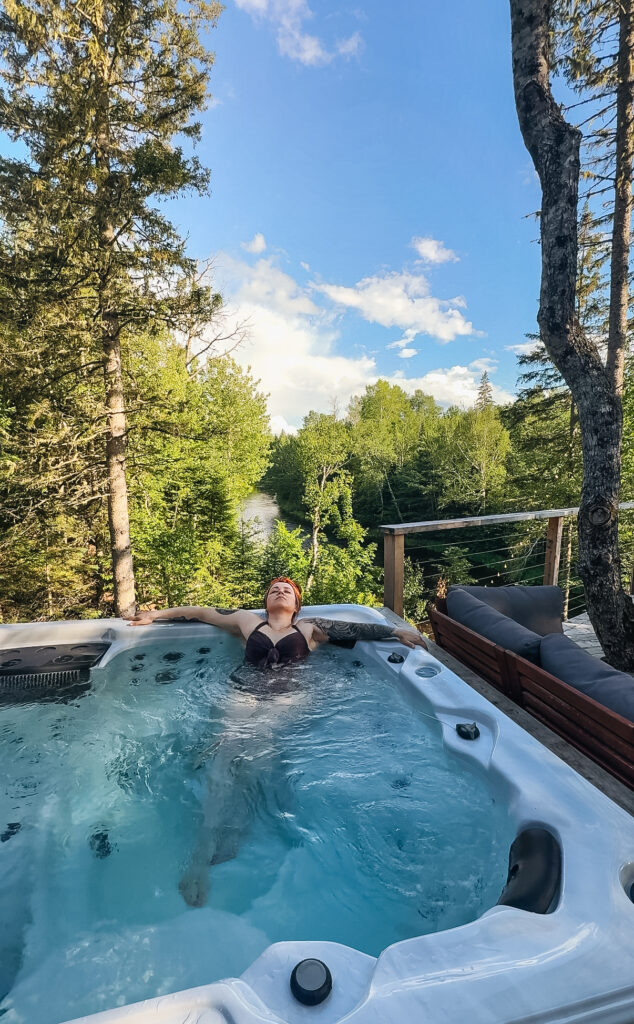 Melissa Danielle in the outdoor hot tub at Eagles Nest, with the river and forest visible in the background on a sunny day
